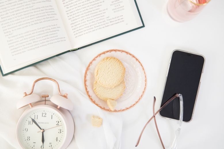 An alarm clock, bowl with cookies, phone and book on a desktop - things to do before 9 am for productivity. 