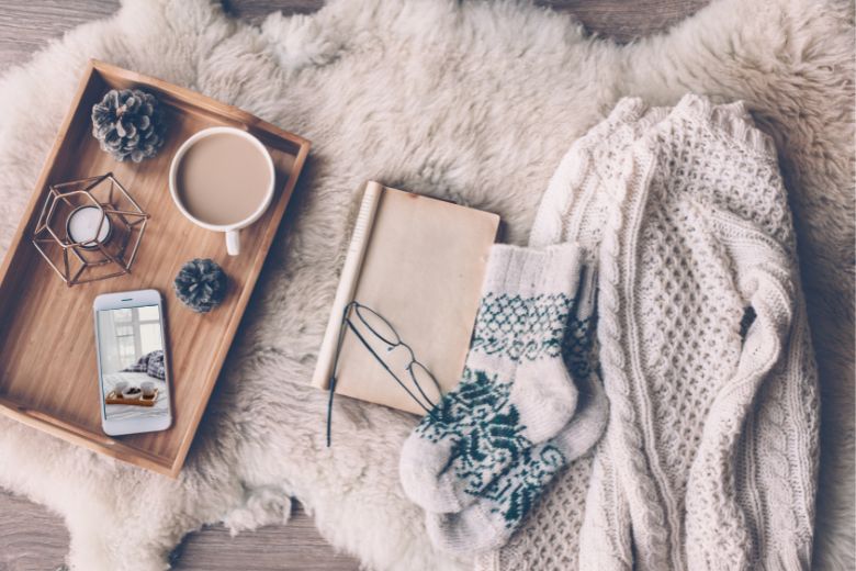 A flat lay in beige of a sweater, socks, book and glasses, and a wood tray with a cup of coffee and an iPhone with a visual - The best winter wallpapers for iPhone lock and home screens. 