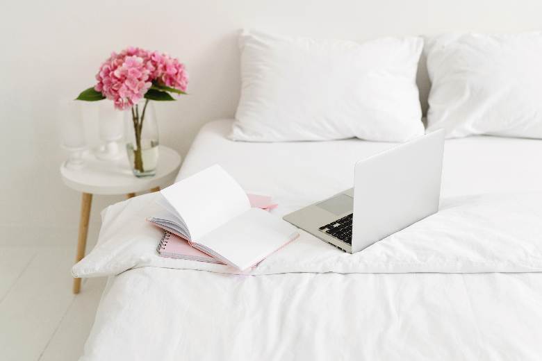 Laptop and notebooks on a white duvet-covered bed - create a blog content calendar. 