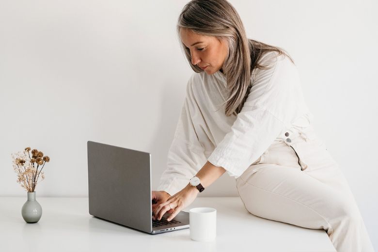 Blog post ideas for midlife bloggers - woman inn her 40s typing on a laptop while sitting on top of a white table. 