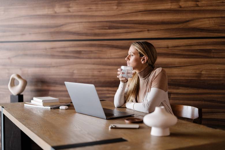 Woman looking through the window while sitting at her laptop, drinking from a cup - how to create an online course to sell.