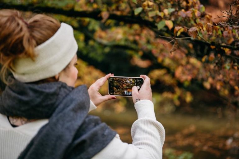 Woman taking a photo of Fall foliage on her phone - Fall hashtags for Instagram.