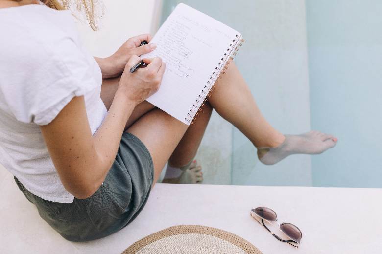 Woman writing in a notebook while sitting by a pool - how to write a travel blog post.