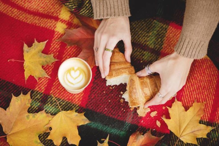 September content ideas - faceless woman breaking open a pastry on a tartan blanket with a cappuccino, outside.
