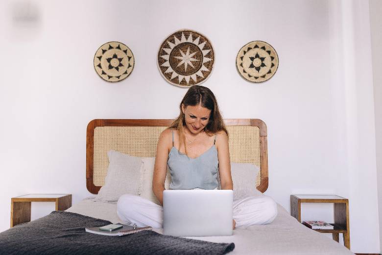 Woman sitting in a hotel room bed using a laptop - travel blog post ideas. 