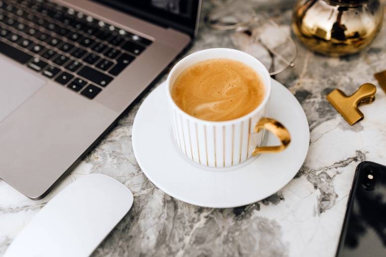 Coffee in a white cup by a laptop and phone - coffee quotes for Instagram posts and captions.