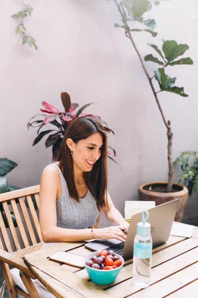 Woman sitting at a table with a laptop and bowl of fruit, typing - profitable blog niches. 