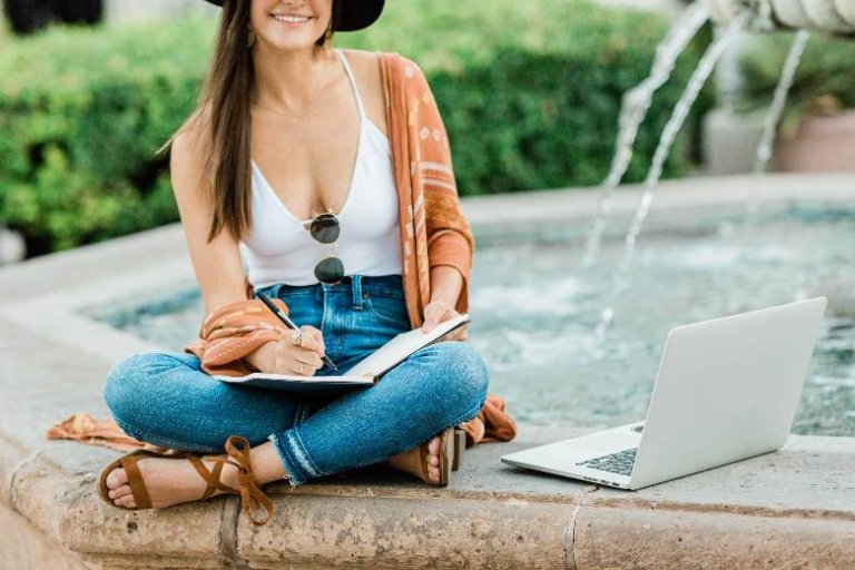 Woman writing in a notebook, while sitting cross-legged on a fountain, with a laptop - how to write a blog post introduction.
