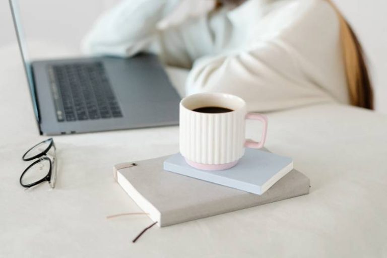 Faceless person leaning on a table by a laptop with a coffee cup and notebook in the foreground - Monday quotes for motivation.